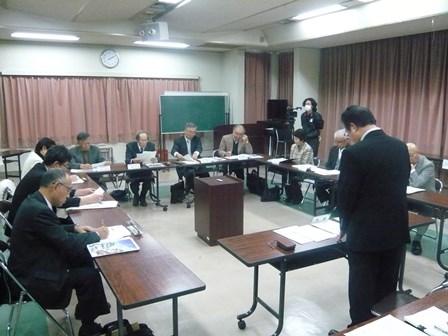 会議室に並んだ茶色いテーブルで前に立つスーツの男性と協議をしている写真