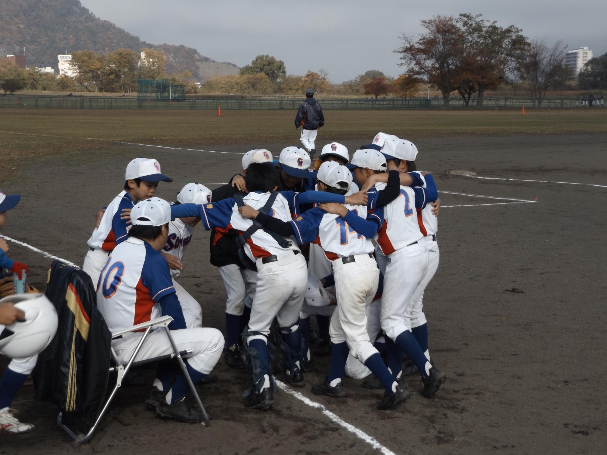 野球のグラウンド上で、ユニフォームを着た子供たちが集まって肩を組んでいる写真