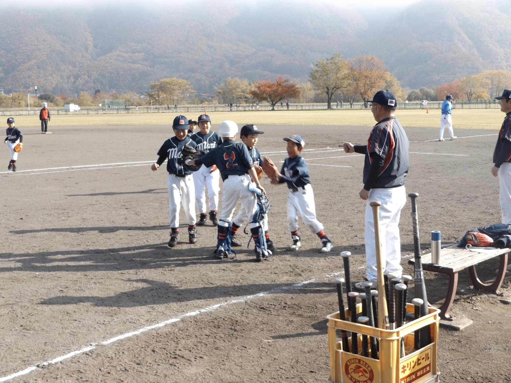 野球のグラウンド上に集まっている子どもたちと、その横で指導している監督の男性の写真
