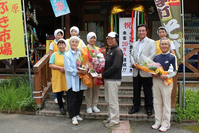 花束を持ったスタッフたちと20万人目の来客者の田中さんが食堂入り口で記念撮影をしている写真