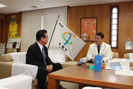 白いスーツの男性が市長と机を挟んで会話している写真