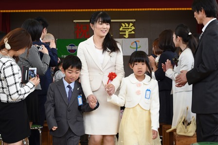 入学式で女性を真ん中に男の子と女の子が手をつないで歩いている写真