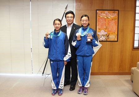 メダルとトロフィーを持ったシンクロ選手の女子小学生の2人の間に市長が立っている写真