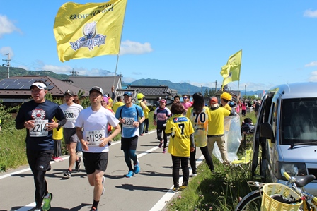 民家が見える道を走るマラソンランナーたちと黄色い旗の写真