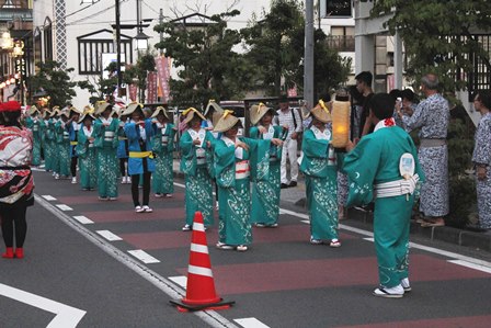 青緑色の着物と笠を被った人たちが夏祭りの路上で並んでいる写真