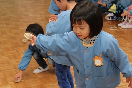青いスモックを着た女の子がおぼつかない手つきで紐を巻いた駒を投げようとしている写真
