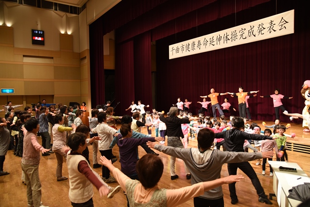 体育館で手をひろげて健康体操を踊る子供たちと高齢者たちの写真