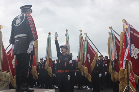 消防ポンプ操法大会での宣誓式で宣誓している団員1名とその後ろに団旗をもって集合する団員達の写真