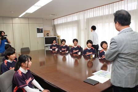 机の両側に着席し、市長と談話するユニフォーム姿の女子生徒達の写真
