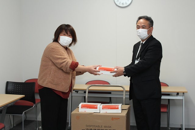 カメラのほうを向きながらマスクの受け渡しをする斉木さんと支援部長の写真