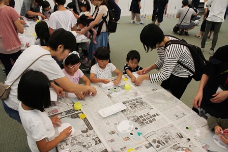 子どもたちが新聞紙を引いた机で大人と一緒に工作をしている様子の写真