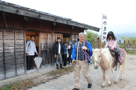 小屋の近くでおじいさんに教わって乗馬体験をしている女の子の写真