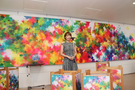 カラフルなアートが飾られた会場内で女性がマイクの前に立っている写真