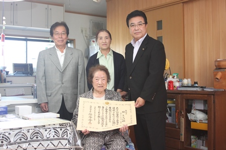 100歳を迎えたおばあさんが賞状をもってリビングで家族と市長と映っている写真