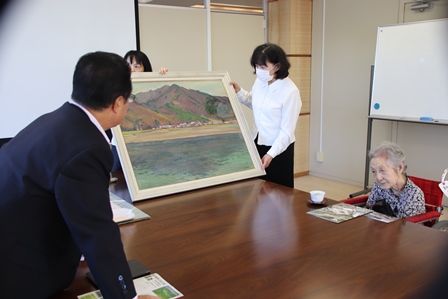 大きい風景画を女性が持ち市長がそれを見ている写真