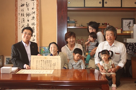 市長と一緒に100歳の賞状を持ったおばあさんが3人の孫と家族と一緒に和室にいる写真