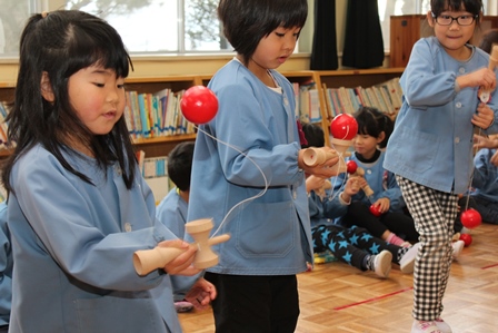 青いスモックを着た女の子の保育園児三人が赤い球のけん玉にチャレンジしている写真