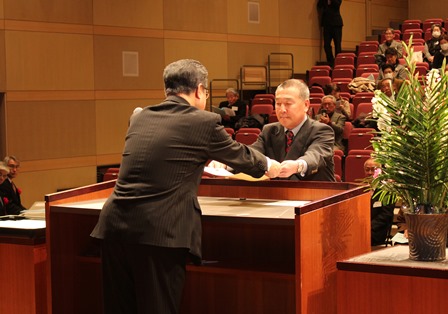 ホールの壇上で市長から卒業証書を受け取る男性の写真