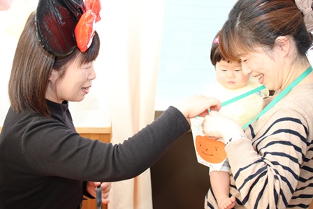 ハロウィンイベントで仮装した女性が親子に首飾りを渡している写真