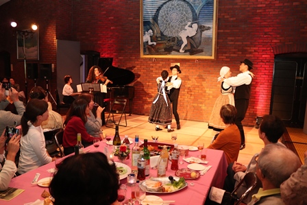 ステージで演奏とダンスが行われる中、食事やワインを楽しんでいる参加者たちの写真