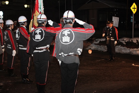警察服姿の岡田市長と、向かい合って敬礼をする消防団員達の写真