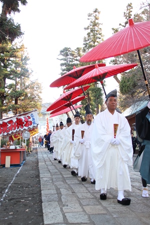 白い装束を着て赤い傘を持った行列が神社の境内を歩いている写真