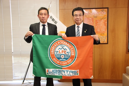 リベルタス千曲FCのロゴが大きくプリントされた旗を、岡田市長と塚口清文監督二人で手に持ちこちらに見せている写真
