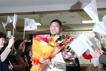 手持ちの紙製の国旗で応援されながら花束を両手に抱えて写真に撮られている中村選手