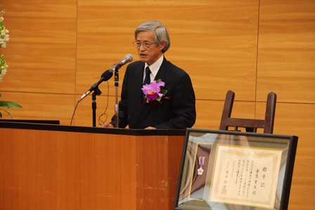 コサージュをつけて演台でスピーチをしている日本画家の男性の写真