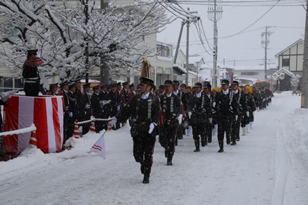 雪道を行進する消防団員と沿道で行進を見ている団員の写真