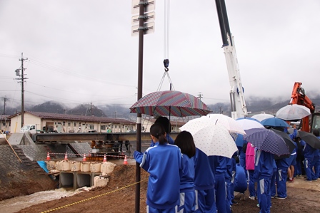 傘を差しながら架設現場の様子を見学する学生らの写真