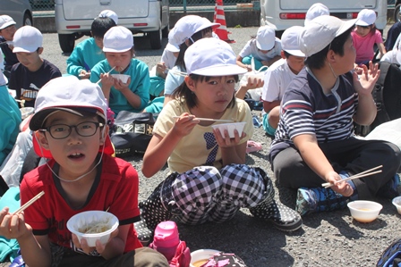 屋外のアスファルトに座り込み、皿を手に食事をしている児童達の写真