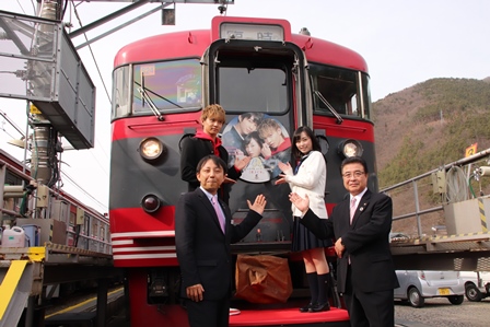 ラッピング電車の前に立ち、市長と主演のおふたりが記念撮影をしている写真