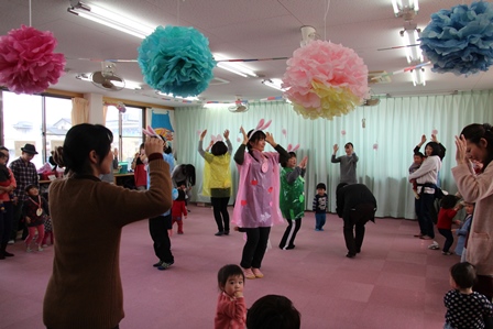 天井からつるされたポンポン飾りと両手を挙げて踊りを踊る親子たちの写真