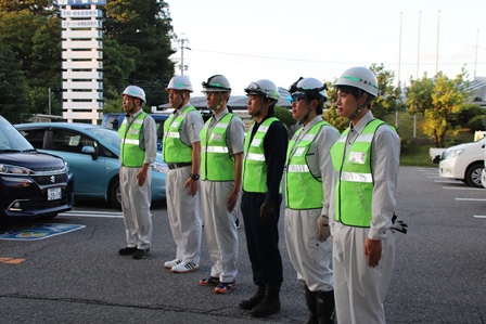 駐車場で緑のチョッキを着た男性職員6人が並んでいる写真