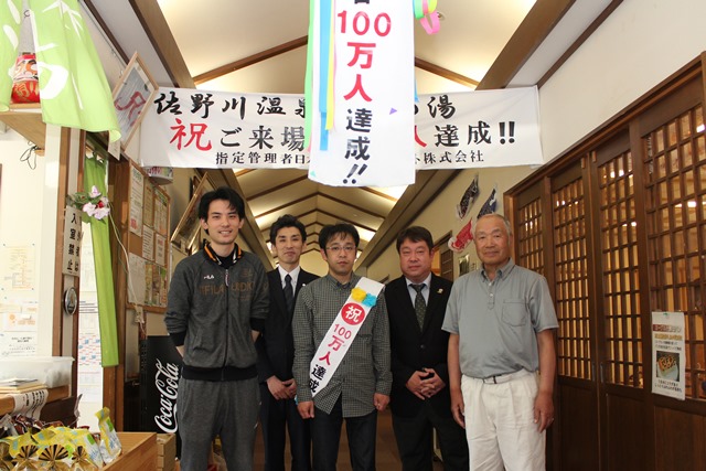 100万人達成！と書かれた垂れ幕とタスキを付けた小林さん、中里さん、宮城さんらが記念撮影をしている写真