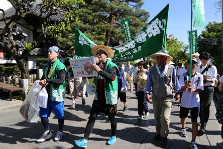 緑色の半被を着た人たちが笑顔で旗やプラカードを持ちながら練り歩く写真