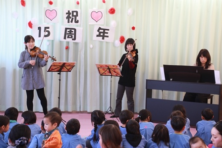 バイオリンを演奏する女性二人とピアノを演奏する女性と演奏を聴く園児たちの写真
