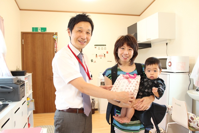 あぷりこっこの施設内で子供を抱いた飯島さんに記念品を手渡している塚田さんの写真