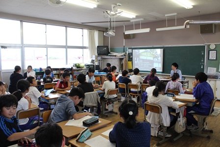 黒板に映る資料を見ながら教室で語り合う先生と生徒の写真