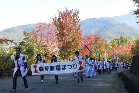 白い衣装を着た人々の行列が紅葉の木々の中の道をパレードする写真