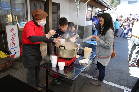 鍋から湯気が立ちのぼる中、味噌汁をプラスチックの容器に注ぎ入れてふるまっている女性の写真