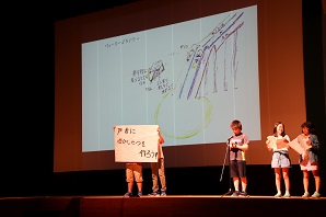 大きなスクリーンのあるステージ上で子供たちが発表をしている写真