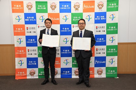 寿高原食品株式会社の水井代表取締役と小川市長が並んで署名した協定書を披露する様子