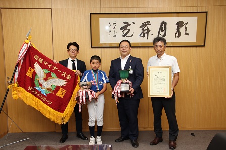 大きな飾り旗やトロフィーなどと一緒に市長と記念撮影をする関田くんの写真