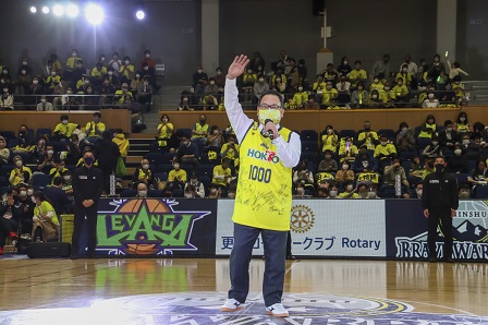 黄色いユニフォームを着た男性が片手をあげて笑顔で挨拶をしている写真