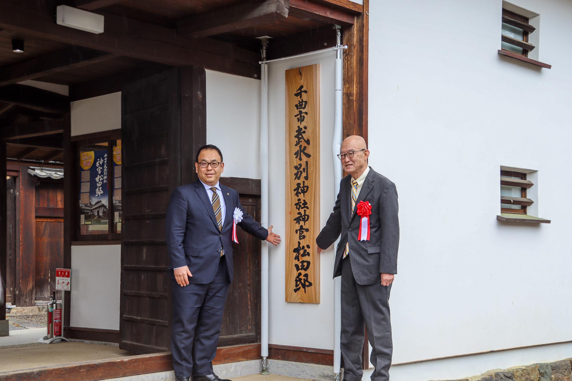 小川修一市長と館名版に揮毫した川村龍洲・驥山館館長が館名板を挟んで記念撮影