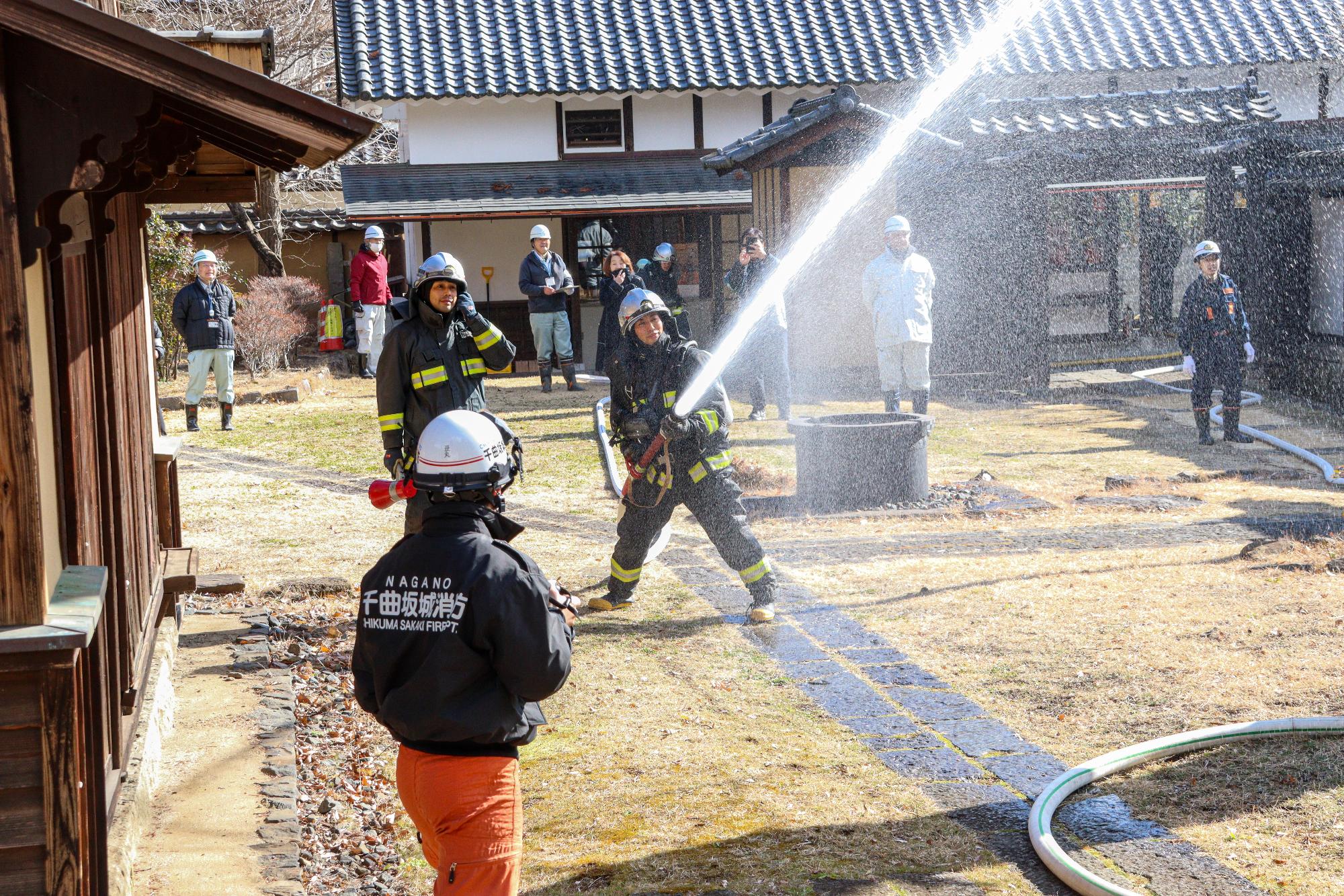 放水訓練で筒先をもって水を出す消防署隊員とその様子を広報で見守る参加者（約10人）