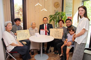 表彰状を手にした峯村さんとご家族の皆さんがカメラに向かって笑顔を見せている写真