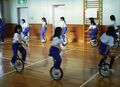 室内で子どもたちが一輪車に乗り、円を描くように列になって走っている写真